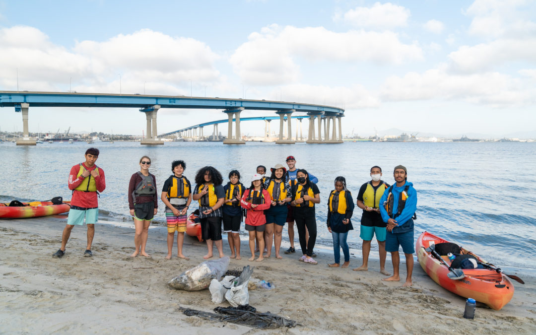 Nine Outdoor Outreach youth participants and four instructors stand together on a small beach with San Diego Bay and Coronado Bridge in the background.