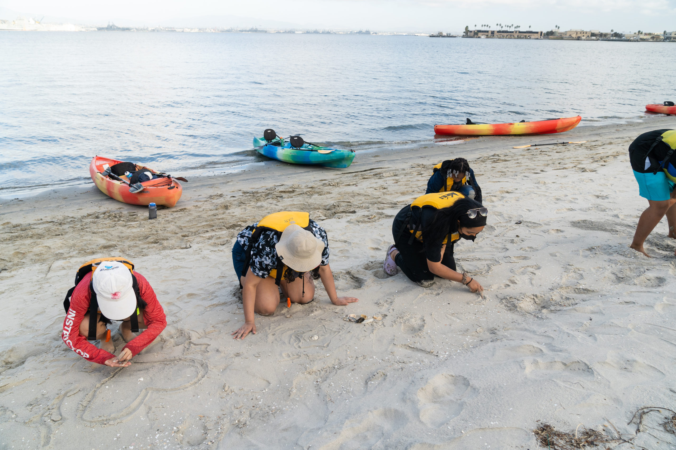 Four Outdoor Outreach youth participants kneeling down on the beach to write and draw on the sand.  